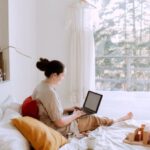 Online Activity - Side view of cute toddler girl sitting on bed barefoot and playing with colorful wooden blocks while mother using laptop in bed enjoying morning coffee