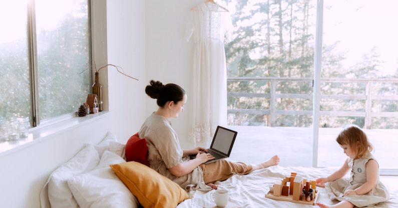 Online Activity - Side view of cute toddler girl sitting on bed barefoot and playing with colorful wooden blocks while mother using laptop in bed enjoying morning coffee